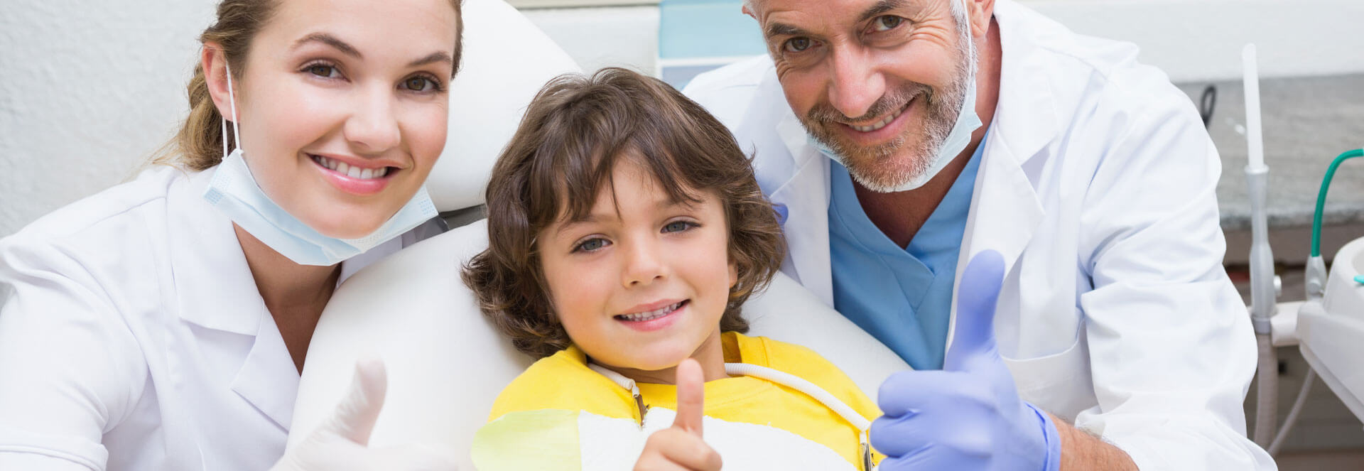 Dental assistant, young child, and dentist all smiling at the camera and showing thumbs up